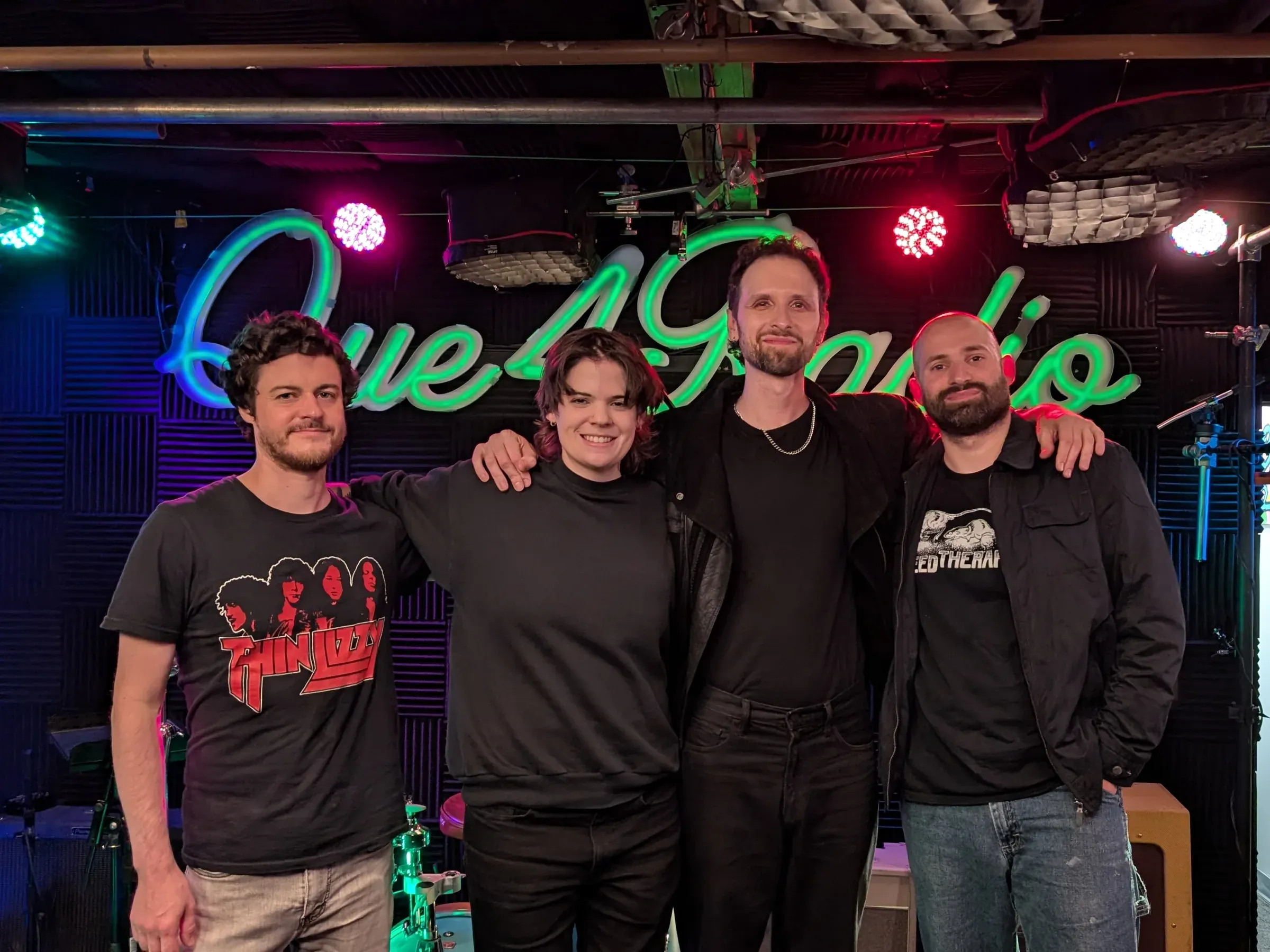 The four members of Pasha & The Pears in front of the Que4 Radio neon sign